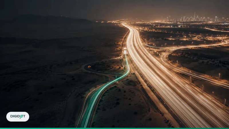 Aerial night view of a desert highway interchange where a broad river of white light trails dwarfs one thin emerald side road, a metaphor for AI referral traffic beside Google organic